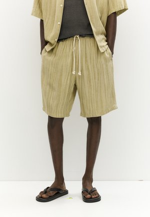 Jasnooliwkowa lniana koszula i szorty w pionowe paski, z elastycznym paskiem w talii i sznurkiem, zestawione z czarnymi klapkami.