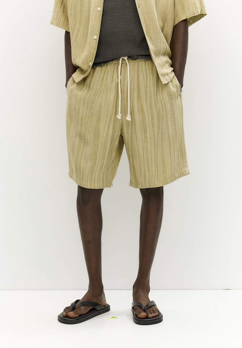 Ensemble en lin olive clair avec des rayures verticales, comprenant une chemise et un short, taille élastique avec cordon de serrage, associé à des tongs noires.