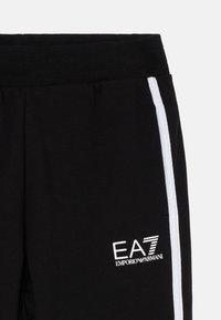 Čierne teplákové nohavice s bielym bočným pruhom, elastickým pásom a bielym logom EA7 Emporio Armani na ľavej nohe. Mäkka textúra tkaniny.