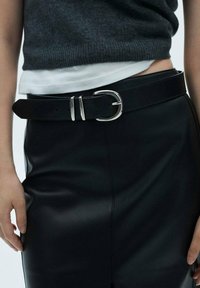 Ceinture en cuir noir avec une grande boucle double en argent, portée sur une jupe en cuir noir, associée à un haut en tricot gris et une chemise blanche.