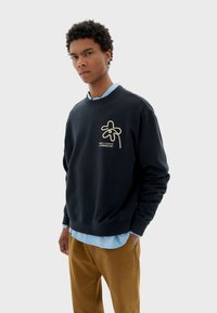 Felpa navy con collo a giro, caratterizzata da una grafica e un testo ricamati bianchi sul fronte. Indossata sopra una camicia azzurra chiara, abbinata a pantaloni marroni.