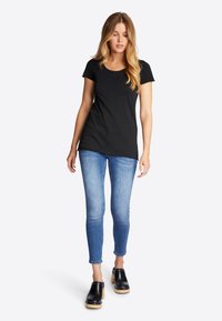 Kvinde med langt blondt hår iført sort T-shirt med korte ærmer, lyseblå skinny jeans og sorte træsko med hæl, stående foran en hvid baggrund.