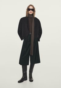 Person wearing oversized black jacket, wide-leg black trousers, tall black boots, brown scarf, brown top, and large dark sunglasses.