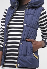 Marinblå pufferväst med avtagbar huva, med dragkedja fram, guldfärgade detaljer och två framfickor. Bärs över en randig tröja.