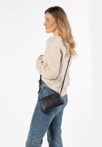 Schwarze Leder-Umhängetasche mit rechteckiger Form, Reißverschluss und verstellbarem Tragegurt, kombiniert mit einem beigen Pullover und blauen Jeans.