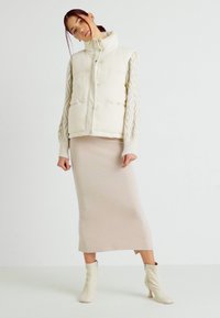 Gilet rembourré en ivoire avec un col montant, associé à une jupe midi crème et des bottes à talons beiges ; accent en tricot sans couture.