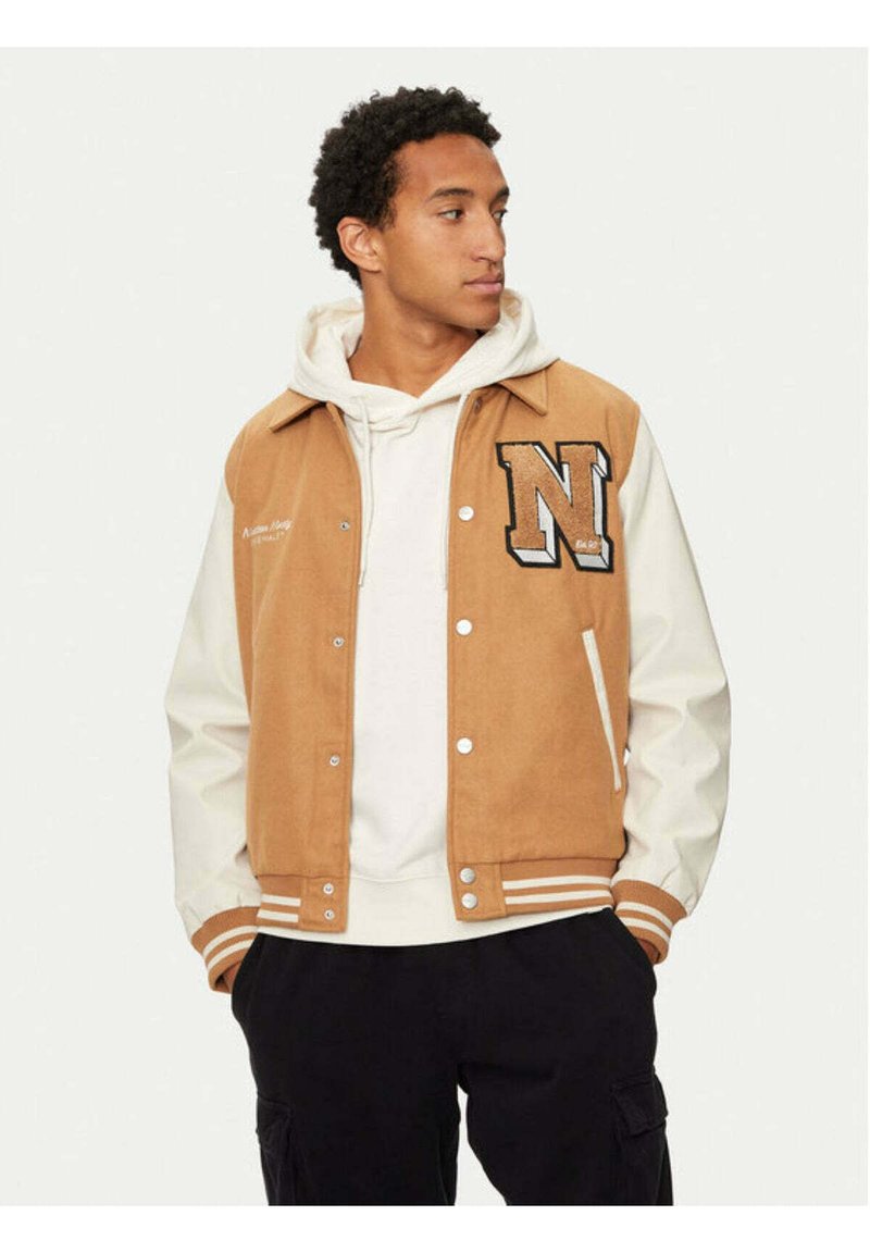Giacca varsity marrone e crema con una toppa "N", bottoni a pressione, polsini a righe e una texture morbida, indossata sopra un hoodie bianco.