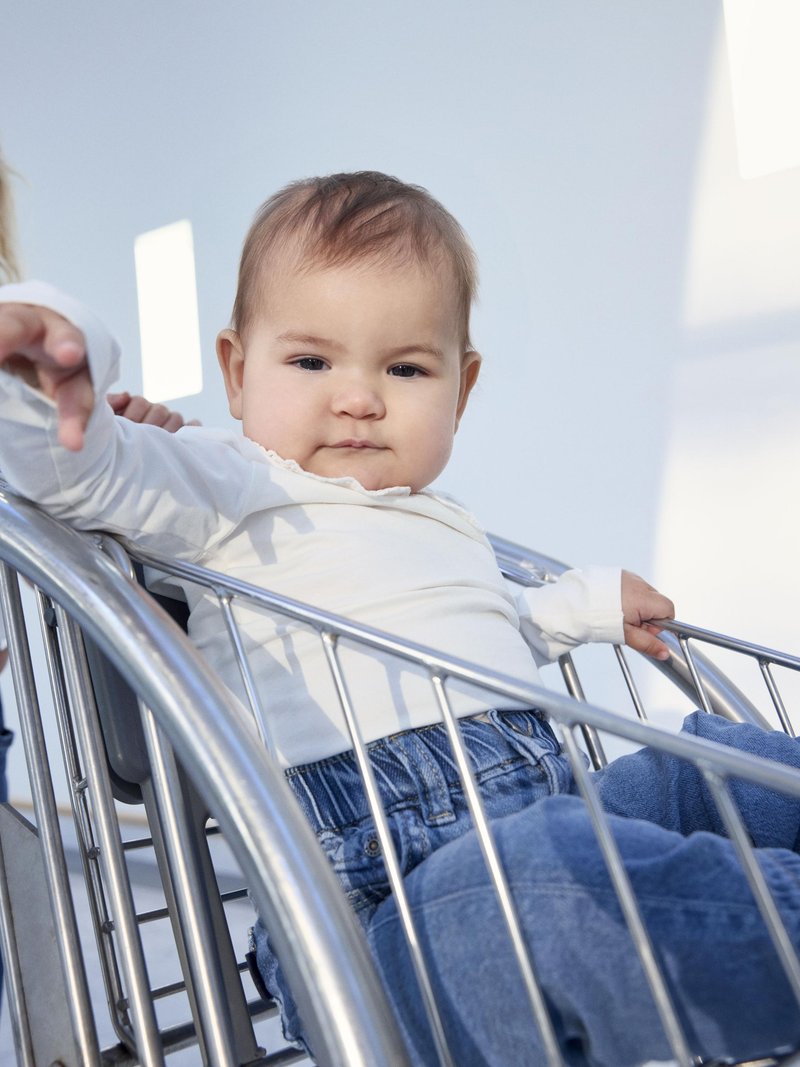 Un bébé est assis dans un chariot de courses en métal argenté. L'enfant porte une chemise blanche à manches longues et un jean en denim bleu avec une texture douce.