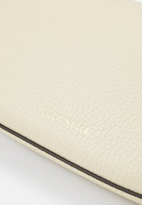 Superficie in pelle testurizzata color crema con la scritta "COCCINELLE" in rilievo con piccole lettere dorate vicino a un bordo cucito.