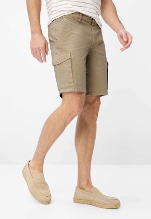 Mand iført beige cargoshorts og lyse slip-on sko, stående mod en ensfarvet hvid baggrund.