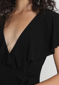 Mujer vistiendo un vestido negro con escote en V pronunciado, mangas cortas con volantes y un lazo en la cintura, con el cabello rizado visible en los hombros.