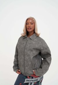 Giacca in ecopelle grigia con colletto e chiusura a zip. Presenta texture sottili e un logo impresso sul lato sinistro.