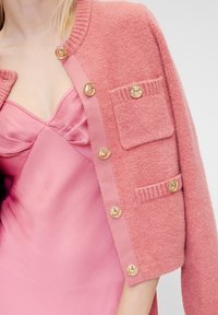 Roza pleteni kardigan sa zlatnim gumbima, ima jedan džep, slojevit preko ružičaste satenske haljine s V-izrezom. Mekana tekstura i kroj prilagođen tijelu.