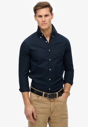 Navy blue button-up shirt with a collar and front pocket, paired with beige trousers and a black belt, featuring a silver watch on wrist.