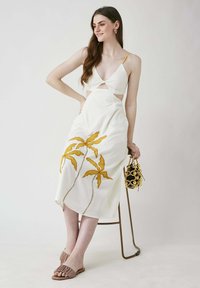 Weißes Kleid mit gelber Palmenblattbestickung, ausgestanztes Design in der Taille, dünne Träger, kombiniert mit beigen Schuhen und einer schwarzen Handtasche mit Perlen.