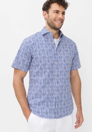 Mann trägt kurzärmliges Hemd mit vertikal blauen und weißen Streifen, steht mit einer Hand in der Tasche und lächelt.