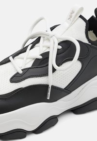 Zapatilla en blanco y negro con tejido de malla, detalles de cuero suave, gruesos cordones blancos y una suela de goma negra texturizada.