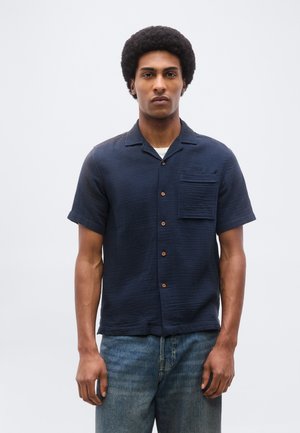 Jonge man met afro kapsel, gekleed in een marineblauw overhemd met korte mouwen en blauwe spijkerbroek, staand tegen een effen lichte achtergrond.