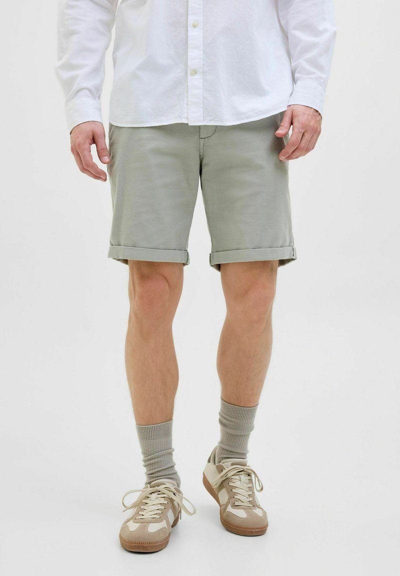 Pantaloni scurți khaki cu manșete răsucite, confecționați dintr-un material moale. Combinați cu teniși bej și șosete gri deschis. Cămașă albă cu butoni purtătă desfăcută.