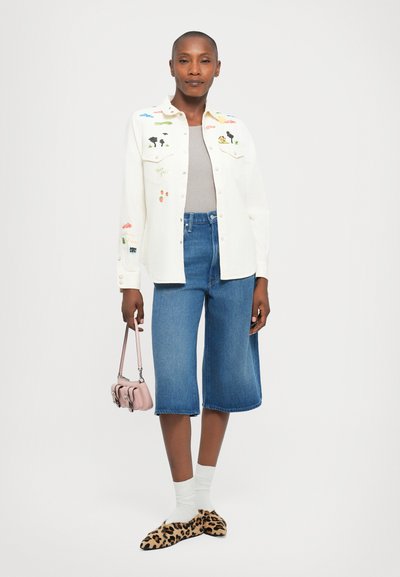 Woman wearing a white embroidered jacket, gray top, knee-length denim shorts, white socks, leopard print shoes, and holding a small pink handbag.