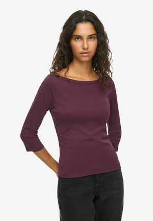Femme aux longs cheveux bouclés portant un haut ajusté violet foncé à manches trois-quarts et un pantalon noir, debout les mains derrière le dos.