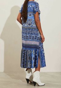 Modrá vzorovaná maxi šaty s krátkými rukávy, s rozšířeným lemem a bočními rozparky. Stylizované s bílými špičatými kotníkovými botami.