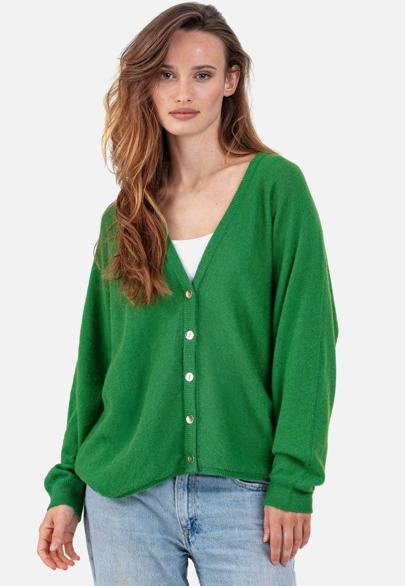Cardigan vert en tissu doux, avec un col en V, des manches longues et des boutons dorés sur le devant. Associé à un haut blanc et un jean en denim.