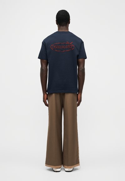 Tmavě modré bavlněné tričko s krátkými rukávy a červeně vyšitou značkou 'VOLTTAIRE' na zádech. Kombinováno s širokými béžovými kalhotami a bílými teniskami.