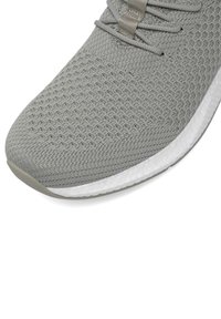 Zapatilla atlética gris con parte superior de malla texturizada, que presenta una entresuela blanca y una suela de goma gris claro. Incluye cordones planos y acentos mínimos.