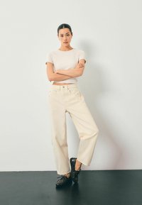 Lichtgekleurde, hoge wijde jeans gecombineerd met een nauwsluitend beige top en donkere instappers tegen een neutrale achtergrond.