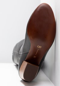 Botte en cuir noir avec surface texturée, bout arrondi et semelle en caoutchouc marron, présentant des marques dorées pour indiquer la taille et les détails du modèle.