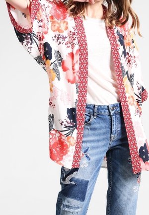 Frau trägt ein weißes, geblümtes Kimono mit rotem Rand über einem weißen Top und blauen, abgenutzten, geflickten Jeans, posiert mit erhobenem Arm.