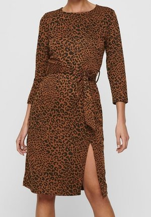 Femme portant une robe marron à motif léopard avec des manches trois-quarts, une ceinture à la taille et une fente devant sur une jambe.