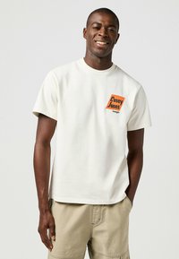 Camiseta de algodón color crema con cuello redondo que presenta un gráfico cuadrado naranja de "Casey Jones" en el pecho, combinada con pantalones cortos cargo beige.