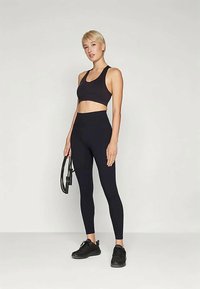 Sujeción deportiva negra y leggings de tiro alto hechos de una tela elástica, con costuras mínimas y una textura suave, combinados con zapatillas deportivas negras y un mat de fitness.