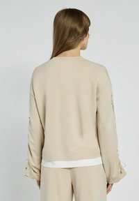 Beżowa bluza z luźnym krojem, z ozdobnymi sznurówkami na rękawach i kontrastowym wykończeniem. Miękki, gładki materiał i minimalistyczny design.