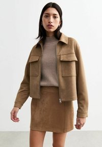 Giacca in suede marrone con due tasche sul petto, chiusura con zip e colletto, abbinata a una gonna marrone coordinata e a un maglione in maglia a trame.