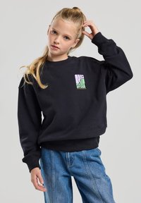 Sweatshirt noir avec des poignets et un ourlet côtelés, arborant un logo coloré sur le côté gauche de la poitrine. Associé à un jean en denim bleu ample.