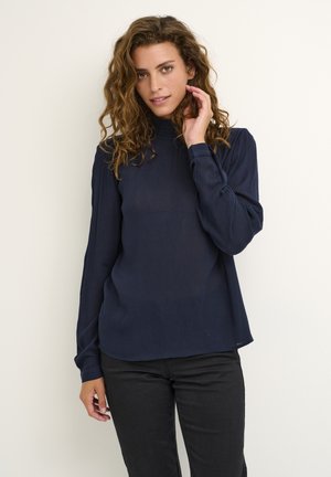 Haut à manches longues bleu marine avec un col montant, texture côtelée et coupe décontractée, associé à un pantalon noir. Design minimaliste et tissu doux.
