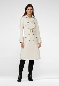 Cream trench coat with a double-breasted design, large buttons, and a belt. Smooth texture and knee-length. Worn over black pants and boots.