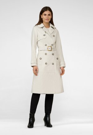 Cream trench coat with a double-breasted design, large buttons, and a belt. Smooth texture and knee-length. Worn over black pants and boots.