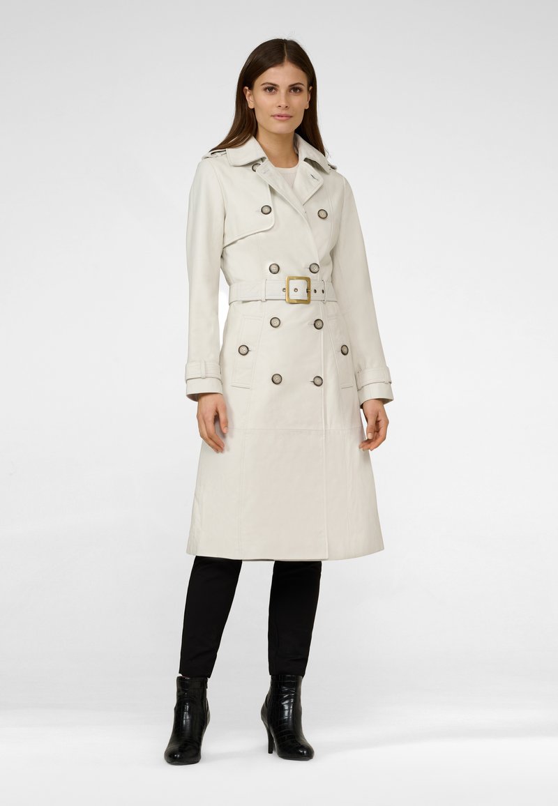 Cream trench coat with a double-breasted design, large buttons, and a belt. Smooth texture and knee-length. Worn over black pants and boots.
