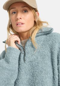 Pullover in pile azzurro chiaro con collo zip, superficie testurizzata e cappuccio. Il colletto e i polsini sono rifiniti con un bordo a contrasto.