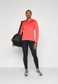 Tætsiddende koralfarvet zip-up hoodie, sorte leggings og grå træningssko. Holder en sort duffelbag med en rund form og strukturstof.