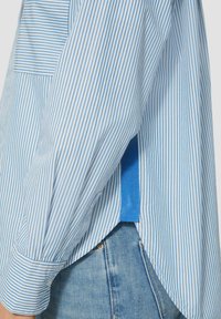 Blauw en witte gestreepte shirt met lange mouwen, voorzien van een afgeronde zoom en een blauwe accent aan de zijkant, gemaakt van lichtgewicht stof.