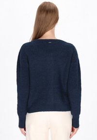 Granatowy sweter wykonany na drutach, o luźnym fasonie, z prążkowaną fakturą i długimi rękawami. Posiada szeroki dekolt oraz niewielki detal z logo z tyłu.