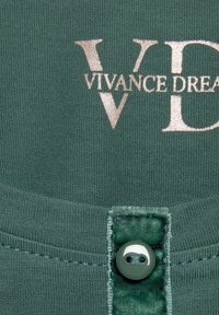 Top in tessuto verde con logo argentato "VIVANCE DREAM" e dettaglio di bottone lucido, con cuciture testurizzate lungo l'orlo.
