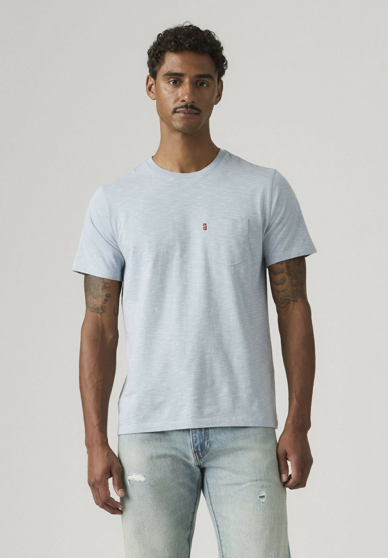 Giovane uomo con capelli ricci e baffi, indossa una T-shirt azzurro chiaro con tasche e jeans strappati, in piedi davanti a uno sfondo semplice.