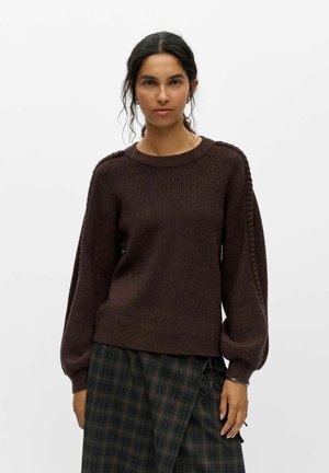 Femme aux cheveux foncés portant un pull en tricot marron avec un détail tressé sur les manches et une jupe écossaise foncée, debout devant un fond blanc uni.