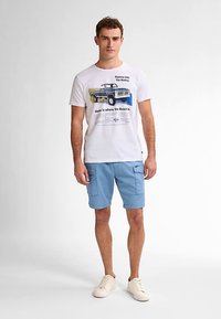 Valkoinen t-paita, jossa on vintage-auton kuvio, yhdistetty vaaleansinisiin shortseihin, joissa on sivutaskut. Malli käyttää beigenvärisiä lenkkareita.